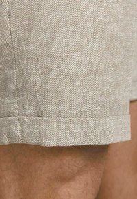 Vaaleanbeiget shorts, joissa on herringbone-kuvio, sileä pinta ja käännetty helma alareunassa, korostaen yksinkertaisia rakentamisdetaljeja.