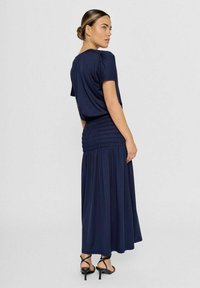 Marineblauwe maxi-jurk met korte mouwen, gerimpelde taille en vloeiende rok. Zacht stof met minimale details. Draagt met zwarte hakken.