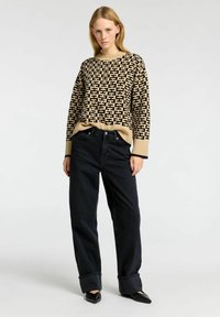 Beige villapaita mustalla geometrisella kuviolla, yhdistettynä löysiin mustiin farkkuihin ja teräväkärkisiin mustiin kenkiin. Villapaidassa on raidalliset hihansuut.