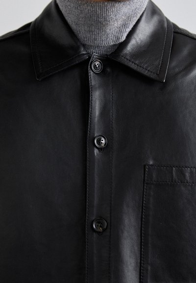 Svart läderskjorta med en slät yta, med en spetskrage, framknäppning och en bröstficka. Mjuk grå polokrage under.