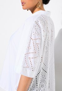 Blusa bianca con dettagli in pizzo ricamato sulle maniche, caratterizzata da una vestibilità morbida e un colletto. Il tessuto testurizzato aggiunge interesse visivo.