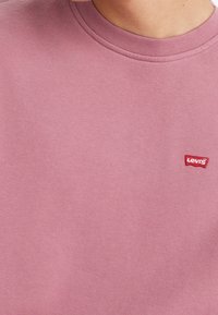 Ljust rosa sweatshirt med rund halsringning och ribbade ärmslut. Har ett litet rött Levi's logotypmärke på vänster sida nära bröstet.