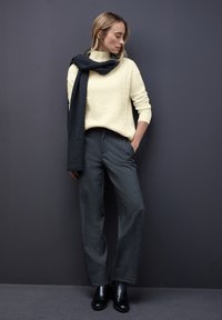 Sweter w kolorze off-white wykonany techniką warkocza, zestawiony z szerokimi spodniami w odcieniu ciemnoszarym, dopełniony ciemnym szalem i czarnymi botkami na kostkę, na tle w ciemnym kolorze.