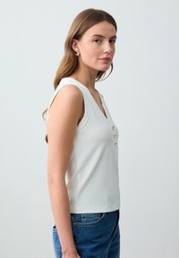 Witte geribde tanktop met een V-hals, voorzien van drie goudkleurige knopen aan de voorkant. Model draagt een blauwe jeans, haar valt soepel over de schouder.
