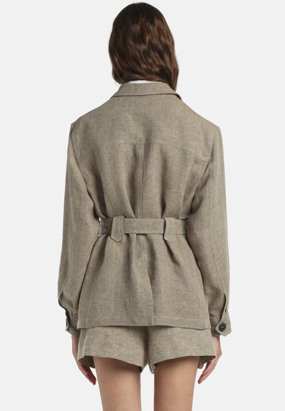 Nő bézs színű, vászon öves dzsekiben, hosszú ujjúval és hozzáillő sorttal, hátulnézetből egyszerű háttér előtt.