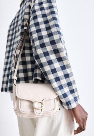 Person trägt ein marineblau-weiß kariertes Hemd und eine kleine weiße Leder-Umhängetasche mit goldener Schnalle auf der rechten Seite.