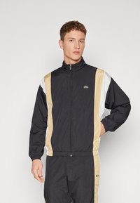 Juodas vėjyje nepralaidus striukė su smėlinės, baltos ir juodos spalvų blokais. Yra užtrauktukas ir elastingos rankogaliai. Lacoste logotipas ant krūtinės.