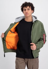Grøn bomberjakke med grå hætte, orange foring, lynlåslommer og rød labeldetalje. Båret over en sort sweatshirt.
