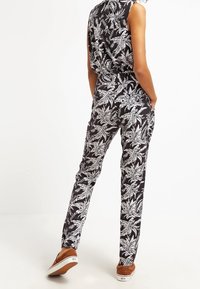 Zwart jumpsuit met een wit palmbladmotief, mouwloos ontwerp, taps toelopende benen en gecombineerd met bruine instapschoenen.