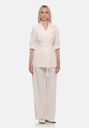 Donna con lunghi capelli biondi che indossa un blazer beige chiaro con cintura, maniche risvoltate, pantaloni a gamba larga abbinati e scarpe bianche.