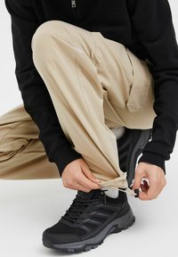 Pantalons cargo beiges avec un ourlet à cordon de serrage, associés à des chaussures de randonnée noires texturées. Sweat à capuche noir avec un col zippé.