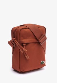 Rustfärgad tygcrossbodyväska med dragkedja, justerbar axelrem, strukturerad yta och logotypaccent. Kompakt rektangulär design.