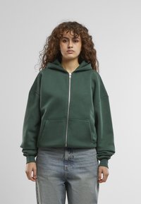 Mørkegrøn zip-up hoodie med kængurulomme, oversized pasform og hætte med snor. Bæres med lyseblå, højtaljede jeans.