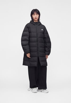Jong persoon draagt een lange zwarte donzen jas met rits, zwarte wijde broek en witte sneakers, staande tegen een effen witte achtergrond.