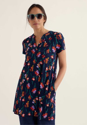 Femeie purtând ochelari de soare teal și o tunică cu mâneci scurte, florală, albastru închis, cu buzunare, stând în fața unui fundal simplu, deschis la culoare.