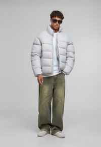 Jachetă puffer gri deschis cu guler înalt, asortată cu pantaloni largi de culoare verde măslinie, cu detalii de cusături curbe. Tenisi albi completează ținuta.