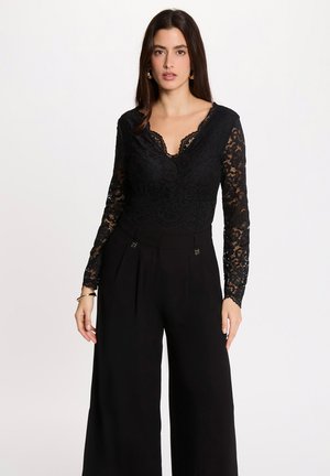 Vrouw draagt een zwarte kanten top met lange mouwen en een gegolfde halslijn en zwarte, hooggetailleerde wijde broek, staand tegen een witte achtergrond.