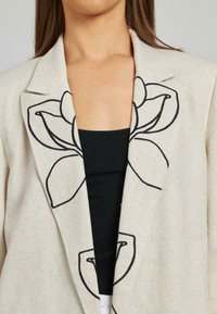 Blazer in lino beige con ricami floreali neri sui risvolti, dotato di colletto a scialle, apertura frontale e superficie in tessuto testurizzato.