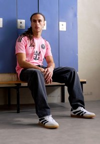 T-shirt rose à manches courtes avec des accents noirs et un logo, associé à un jean foncé et des baskets blanches avec des rayures noires et une semelle en caoutchouc.