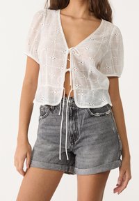 Mujer vistiendo una blusa corta de encaje blanco con mangas cortas atada al frente, combinada con pantalones cortos de mezclilla gris con dobladillo enrollado.