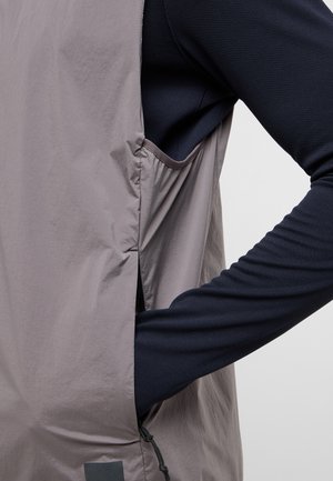 Strato esterno grigio leggero con una texture morbida, dotato di una tasca laterale e uno strato di base aderente a maniche lunghe in blu navy sotto.