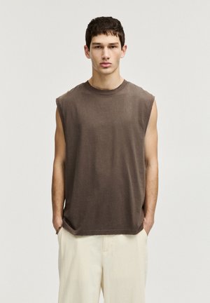 T-shirt marrone senza maniche realizzato in tessuto morbido, con scollo rotondo e bordi grezzi, abbinato a pantaloni chiari e larghi.