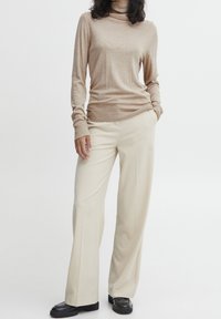 Maglione a collo alto beige in morbido materiale lavorato a maglia, abbinato a pantaloni a gamba larga crema. Scarpe nere completano l'outfit.