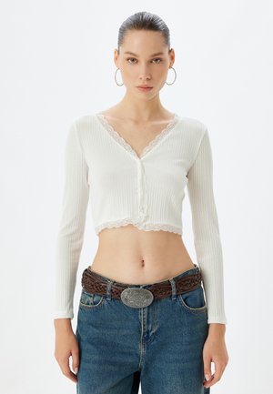Witte geribbelde lange mouwen cropped top met kantafwerking bij de halslijn en zoom, gecombineerd met high-waisted blauwe denim jeans en een bruine gevlochten riem.
