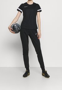 Svart Nike sportkläder-set som består av en kortärmad topp med vita detaljer, slim-fit byxor och svarta fotbollsskor som håller en svart fotboll.