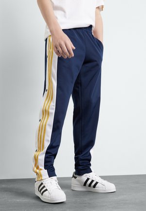 Personne portant un pantalon de survêtement bleu marine avec des bandes latérales blanches et jaunes, et des baskets blanches à rayures noires, debout sur un sol gris.