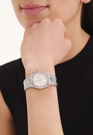 Woman wearing a silver metal band watch with a round, light-colored face and subtle Roman numerals, resting her chin on her hand.