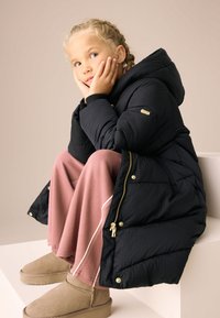 Doudoune noire à capuche, avec une texture matelassée et des accessoires dorés, portée sur un pantalon large rose et des bottines beige.