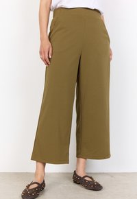 Person wearing olive green wide-leg cropped pants and dark brown studded Mary Jane flats standing on a light floor.