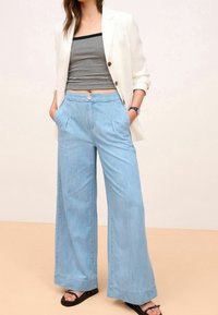 Pantalon large bleu clair en denim doux, doté d'une fermeture à boutons, de deux poches avant et de plis subtils le long des jambes.