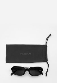 Svarta rektangulära solglasögon med mörka linser och tjocka bågar, placerade ovanför en svart tygpåse med texten "PULL&BEAR."