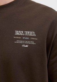 Brown cotton sweatshirt featuring white printed text: "ESS. WRD." and "Worldwide STUDIO Collection" with additional details below.