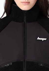Giacca nera con zip realizzata in tessuto felpato e liscio, colletto alto e due tasche con zip. Presenta un logo sul lato sinistro.