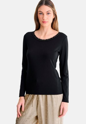 Vrouw draagt een zwart shirt met lange mouwen en kant aan de halslijn en lichtbeige broek met patroon, staande tegen een effen witte achtergrond.