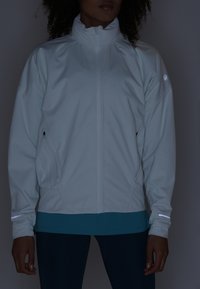 Personne portant une veste zippée gris clair avec des bandes réfléchissantes sur les manches et un pantalon foncé, debout devant un fond sombre.