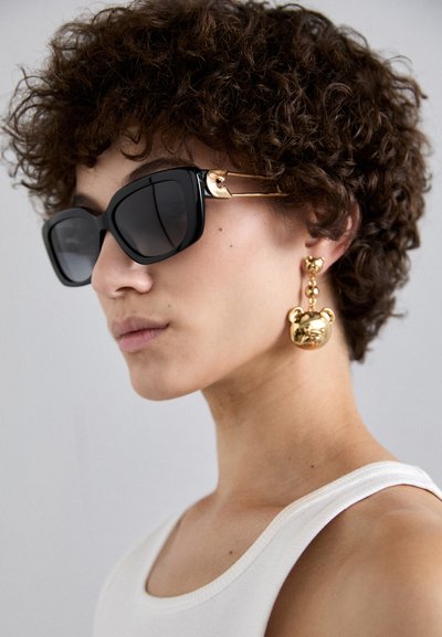 Lunettes de soleil carrées noires surdimensionnées avec garnitures dorées ; boucles d'oreilles assorties en forme d'ours doré. Le modèle porte un débardeur blanc, affichant un look minimaliste.