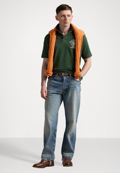 Tricou polo verde cu logo brodat, pulover portocaliu din tricot cu fire întinse pe umeri, blugi albaştri drepti, pantofi din piele maro.