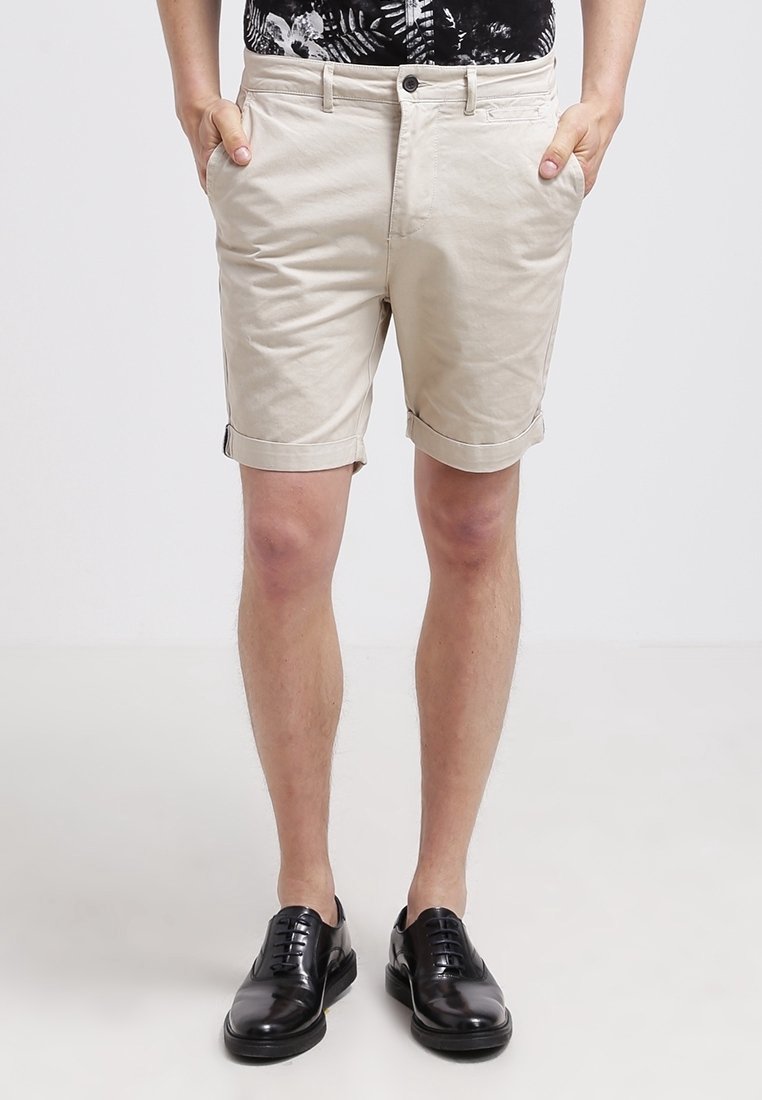 Homme portant un short beige retroussé, des chaussures habillées noires et une chemise à motif noir et blanc, les mains dans les poches.