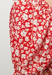 Roter Stoff mit einem floralen Muster, das kleine weiße Blumen zeigt. Das Design ist leicht und hat eine geraffte Textur entlang der Kanten.