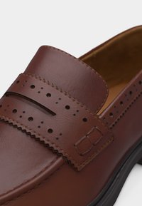 Καφέ δερμάτινο μοκασίνι με διακοσμητικές διατρήσεις και λεπτομέρειες με κυματιστό άκρο, με μαύρη σόλα και λείο φινίρισμα.