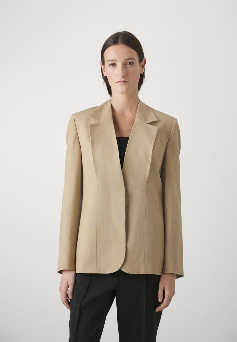 Beige maßgeschneiderter Blazer mit breiten Revers, strukturierten Schultern und glatter Textur. Getragen über einem schwarzen Top, kombiniert mit schwarzen Hosen.