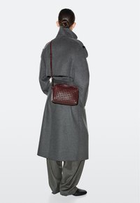 Person mit Dutt, trägt einen langen grauen Mantel, weite graue Hosen, schwarze Schuhe und eine burgunderrote, gewebte Ledertasche mit Schulterriemen.