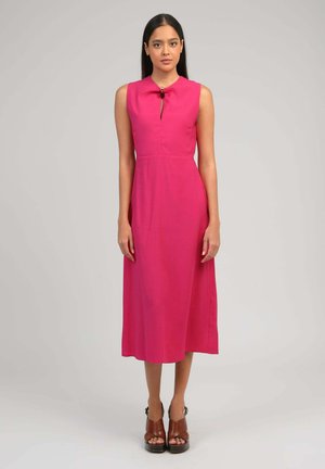 Femme debout portant une robe midi rose vif sans manches avec une ouverture en trou de serrure à l'avant et des talons compensés marron à bouts ouverts sur fond gris clair.