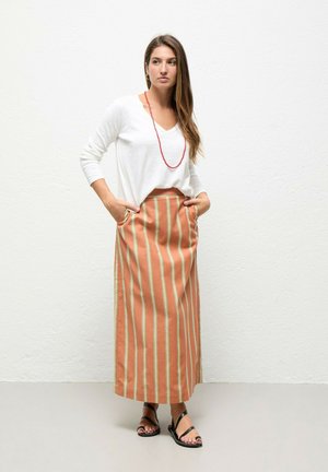 Mujer con una camiseta blanca de manga larga, falda de rayas verticales naranja y beige con bolsillos, sandalias negras y un collar de cuentas rojo, de pie con las manos en los bolsillos.