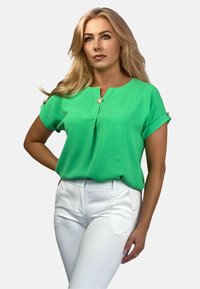 Zielona bluzka z krótkim rękawem, okrągłym dekoltem i dekoracyjnym guzikiem. Noszona z białymi, dopasowanymi spodniami z teksturowanej tkaniny.