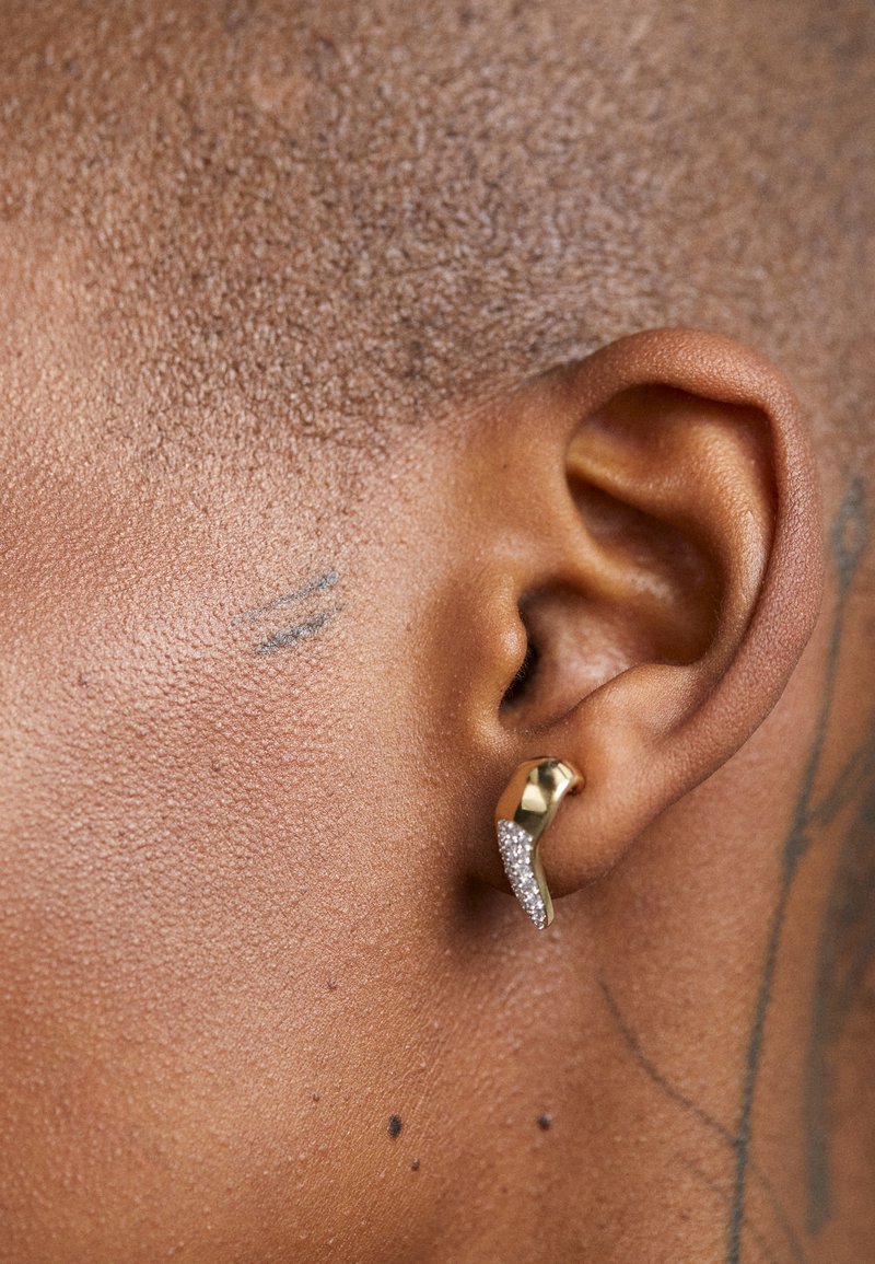 Cercei aurii cu un design unduit; o parte prezintă mici pietre semiprețioase clare, evidențiind textura și contrastul față de piele.
